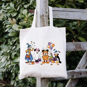 May include: Cream canvas tote bag with a Halloween design. The illustration shows cartoon characters in costumes, including a witch, ghost, and pumpkin. The design also features sweets, balloons, and bats.
