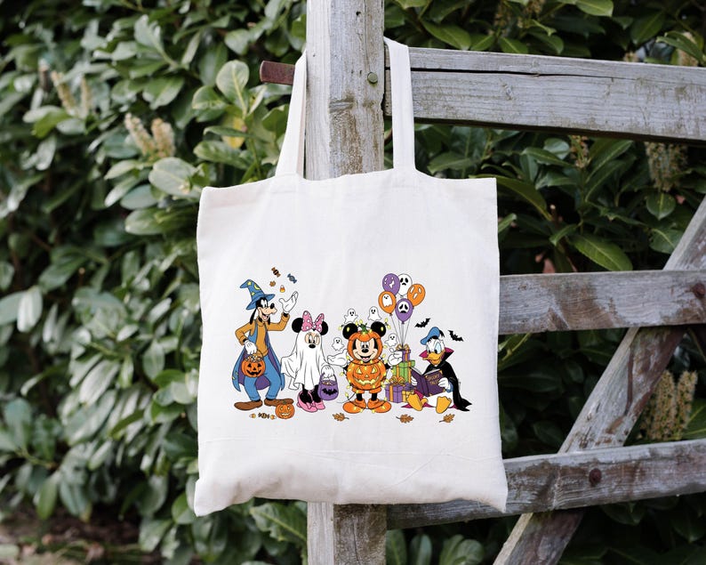 May include: Cream canvas tote bag with a Halloween design. The illustration shows cartoon characters in costumes, including a witch, ghost, and pumpkin. The design also features sweets, balloons, and bats.