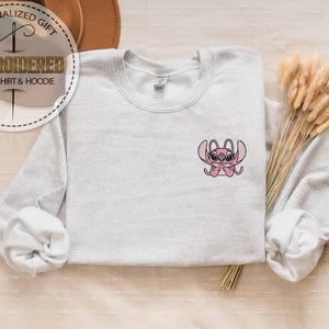 May include: A light gray sweatshirt with a small embroidered pink cartoon character on the chest. The sweatshirt has long sleeves and a crew neck. The text "PERSONALIZED GIFT" and "EMBROIDERED SWEATSHIRT & HOODIE" are in a circular design in the upper left corner.