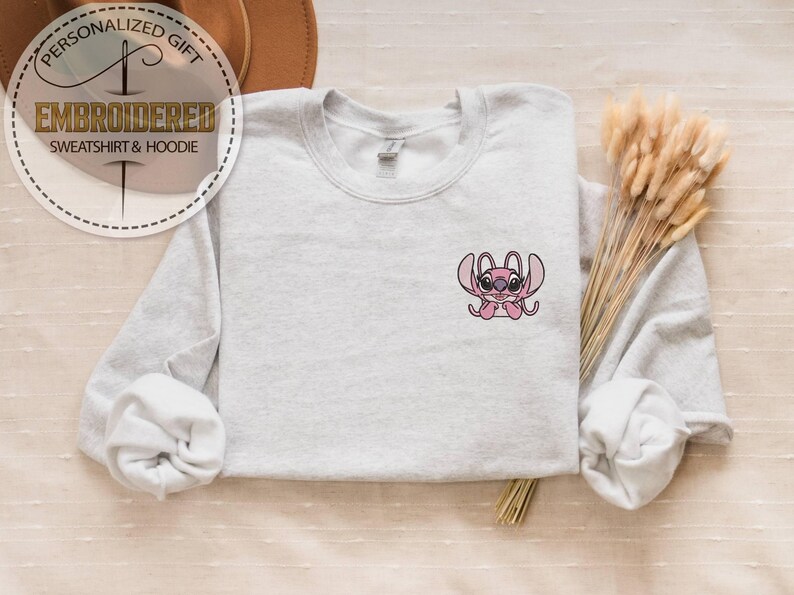 May include: A light gray sweatshirt with a small embroidered pink cartoon character on the chest. The sweatshirt has long sleeves and a crew neck. The text "PERSONALIZED GIFT" and "EMBROIDERED SWEATSHIRT & HOODIE" are in a circular design in the upper left corner.