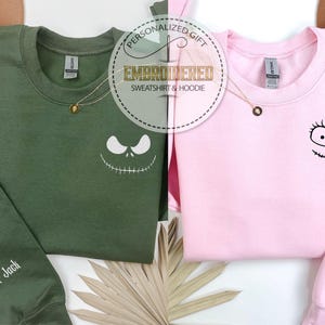 May include: Two sweatshirts, one olive green and one pink, with embroidered character faces. The green sweatshirt has "Her Jack" on the sleeve, and the pink one has "His Sally." Both have gold necklaces and the text "EMBROIDERED SWEATSHIRT & HOODIE".