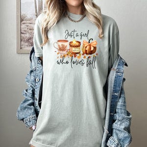 K&ouml;nnte beinhalten: Hellgr&uuml;nes T-Shirt mit dem Text "Just a girl who loves fall". Das Design zeigt eine Kaffeetasse, eine Kerze und einen K&uuml;rbis in Herbstfarben. Ein l&auml;ssiges, saisonales Kleidungsst&uuml;ck.