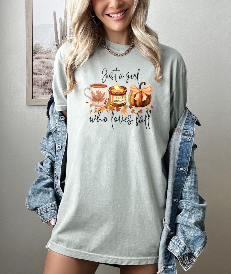 K&ouml;nnte beinhalten: Hellgr&uuml;nes T-Shirt mit dem Text "Just a girl who loves fall". Das Design zeigt eine Kaffeetasse, eine Kerze und einen K&uuml;rbis in Herbstfarben. Ein l&auml;ssiges, saisonales Kleidungsst&uuml;ck.