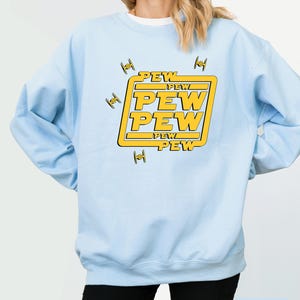 May include: Light blue sweatshirt with a yellow graphic that says "PEW PEW PEW" in a bold font. The design includes small, stylized spaceship illustrations. The sweatshirt is a solid light blue color.