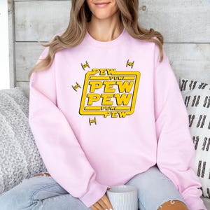 May include: A light pink sweatshirt with a yellow and white graphic that says "PEW PEW PEW" in a bold font. The graphic is surrounded by yellow squares and small black fighter jet illustrations.