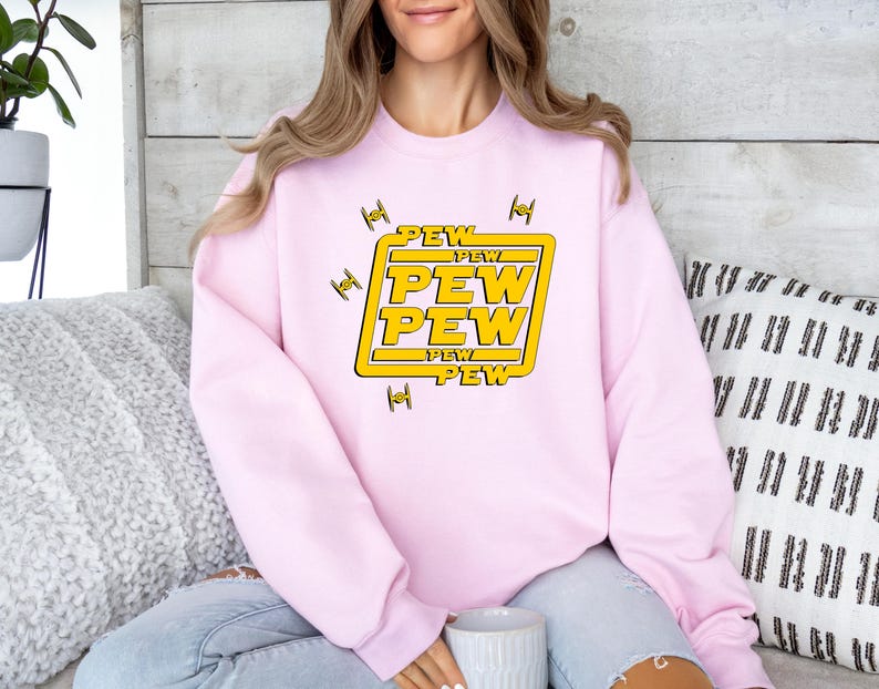 May include: A light pink sweatshirt with a yellow and white graphic that says "PEW PEW PEW" in a bold font. The graphic is surrounded by yellow squares and small black fighter jet illustrations.