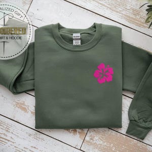 May include: An olive green sweatshirt with a bright pink hibiscus flower design. The sweatshirt has long sleeves and a crew neck. The image also includes text that says "PERSONALIZED GIFT" and "EMBROIDERED SWEATSHIRT & HOODIE".
