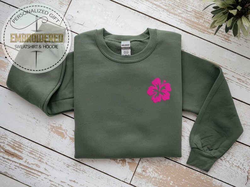 May include: An olive green sweatshirt with a bright pink hibiscus flower design. The sweatshirt has long sleeves and a crew neck. The image also includes text that says "PERSONALIZED GIFT" and "EMBROIDERED SWEATSHIRT & HOODIE".