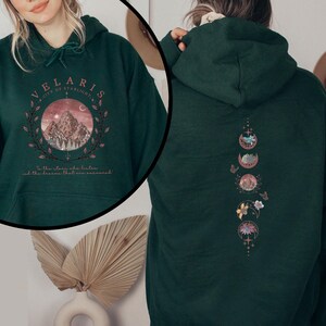 May include: Dark green hoodie with a front graphic featuring a mountain scene within a circle, the text "VELARIS CITY OF STARLIGHT", and the quote "To the stars who listen and the dreams that are answered." The back has a vertical design of moon phases.