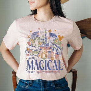 Puede incluir: Camiseta rosa claro con un dise&ntilde;o gr&aacute;fico colorido que presenta un castillo, fuegos artificiales y otros elementos del parque. El texto "The Most Magical Place on Earth" est&aacute; impreso debajo en azul marino.