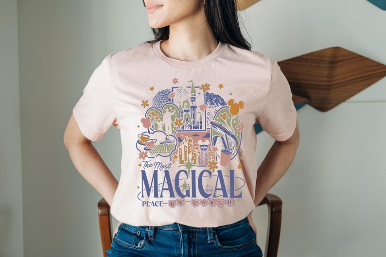 Puede incluir: Camiseta rosa claro con un dise&ntilde;o gr&aacute;fico colorido que presenta un castillo, fuegos artificiales y otros elementos del parque. El texto "The Most Magical Place on Earth" est&aacute; impreso debajo en azul marino.
