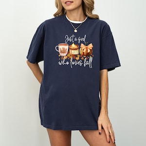 K&ouml;nnte beinhalten: Marineblaues T-Shirt mit einem herbstlichen Motiv. Das Design zeigt eine Tasse, eine Kerze und einen K&uuml;rbis mit Schleife sowie den Text "Just a girl who loves fall".