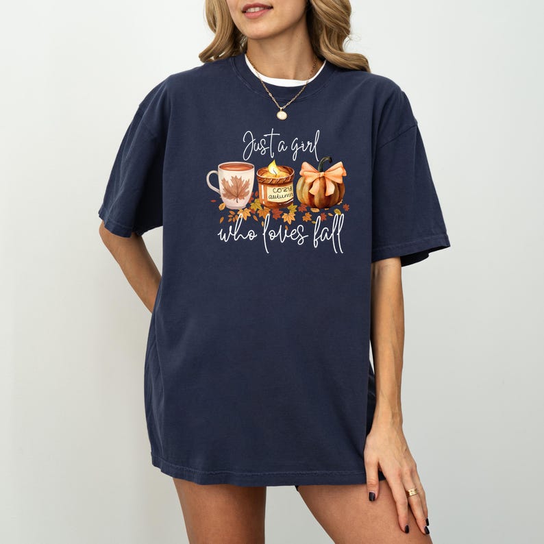 K&ouml;nnte beinhalten: Marineblaues T-Shirt mit einem herbstlichen Motiv. Das Design zeigt eine Tasse, eine Kerze und einen K&uuml;rbis mit Schleife sowie den Text "Just a girl who loves fall".
