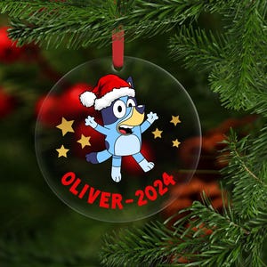 May include: A round, clear acrylic Christmas ornament featuring a cartoon blue dog wearing a red and white Santa hat, surrounded by yellow stars. The text OLIVER-2024 is printed in red at the bottom. A red ribbon is attached to the top for hanging.