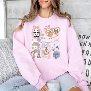 May include: A light pink sweatshirt featuring Star Wars themed illustrations. The design includes a Stormtrooper with a pink bow, a lightsaber, and various characters. The words "STAR WARS" are also printed on the sweatshirt.