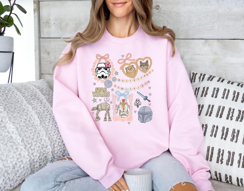 May include: A light pink sweatshirt featuring Star Wars themed illustrations. The design includes a Stormtrooper with a pink bow, a lightsaber, and various characters. The words "STAR WARS" are also printed on the sweatshirt.