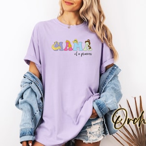 K&ouml;nnte beinhalten: Ein lavendelfarbenes T-Shirt mit einer Grafik des Wortes "Mama", das mit Disney-Prinzessinnen-Charakteren ausgeschrieben ist. Der Text "of a princess" befindet sich unter der Grafik.