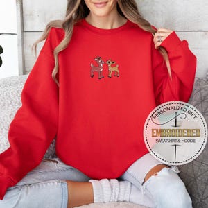 Peut inclure: Sweat-shirt rouge avec un motif brod&eacute; blanc de deux rennes. Le sweat-shirt a un col rond et des manches longues. Le texte "Cadeau personnalis&eacute; Sweat-shirt et sweat &agrave; capuche brod&eacute;" est sur un cercle blanc avec un contour dor&eacute;.