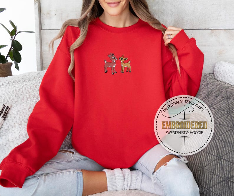 Peut inclure: Sweat-shirt rouge avec un motif brod&eacute; blanc de deux rennes. Le sweat-shirt a un col rond et des manches longues. Le texte "Cadeau personnalis&eacute; Sweat-shirt et sweat &agrave; capuche brod&eacute;" est sur un cercle blanc avec un contour dor&eacute;.