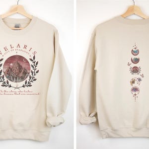 May include: Beige sweatshirt with a front graphic featuring the text "VELARIS CITY OF STARLIGHT" above a mountain scene within a laurel wreath. The back displays a vertical design of moon phases. The text "In the stars who listen and the dreams that are answered" is below the front graphic.