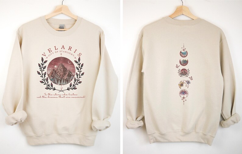 May include: Beige sweatshirt with a front graphic featuring the text "VELARIS CITY OF STARLIGHT" above a mountain scene within a laurel wreath. The back displays a vertical design of moon phases. The text "In the stars who listen and the dreams that are answered" is below the front graphic.