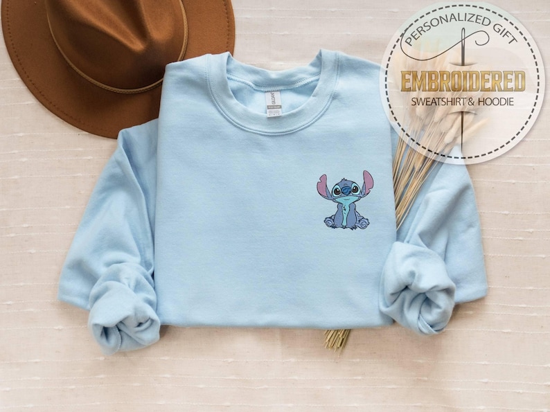 May include: A light blue sweatshirt with a small embroidered image of the Disney character Stitch. The sweatshirt has long sleeves and a crew neck. A brown hat is visible in the background. The text "PERSONALIZED GIFT EMBROIDERED SWEATSHIRT & HOODIE" is printed on a circular sticker.