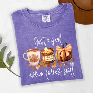 K&ouml;nnte beinhalten: Ein lilafarbenes T-Shirt mit dem Text "Just a girl who loves fall". Die Grafik zeigt eine Tasse, eine Kerze und einen K&uuml;rbis. Das Shirt ist von der Marke Comfort Colors.