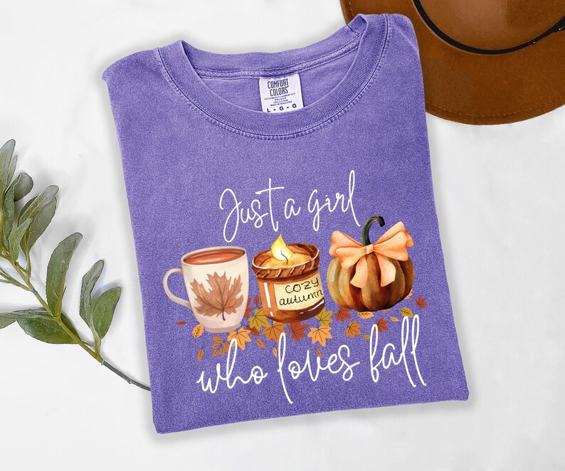 K&ouml;nnte beinhalten: Ein lilafarbenes T-Shirt mit dem Text "Just a girl who loves fall". Die Grafik zeigt eine Tasse, eine Kerze und einen K&uuml;rbis. Das Shirt ist von der Marke Comfort Colors.