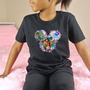 Puede incluir: Camiseta negra con un dise&ntilde;o gr&aacute;fico colorido. El dise&ntilde;o tiene forma de orejas de Mickey Mouse y representa una escena tropical con personajes. La camiseta est&aacute; hecha de un material suave.