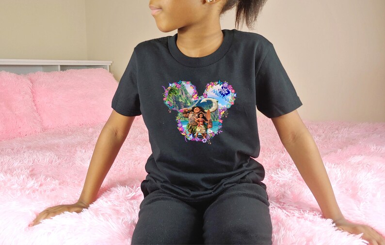 Puede incluir: Camiseta negra con un dise&ntilde;o gr&aacute;fico colorido. El dise&ntilde;o tiene forma de orejas de Mickey Mouse y representa una escena tropical con personajes. La camiseta est&aacute; hecha de un material suave.