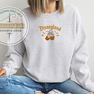 Pode incluir: Um moletom cinza claro com a palavra "Disneyland" em escrita laranja, juntamente com um gr&aacute;fico de ab&oacute;boras e morcegos. O moletom tamb&eacute;m apresenta o texto "EST. 1955". A imagem inclui um logotipo circular que diz "Presente Personalizado Moletom e Moletom com Capuz Bordado".
