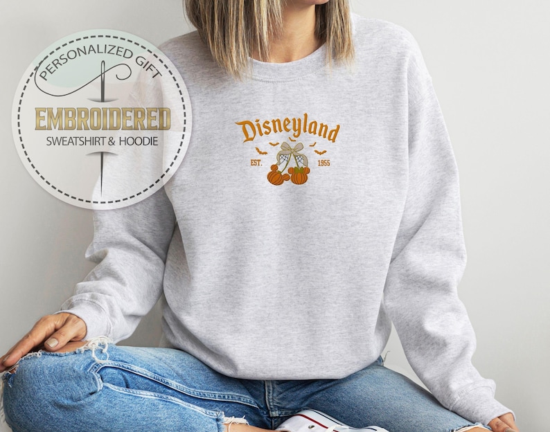 Pode incluir: Um moletom cinza claro com a palavra "Disneyland" em escrita laranja, juntamente com um gr&aacute;fico de ab&oacute;boras e morcegos. O moletom tamb&eacute;m apresenta o texto "EST. 1955". A imagem inclui um logotipo circular que diz "Presente Personalizado Moletom e Moletom com Capuz Bordado".