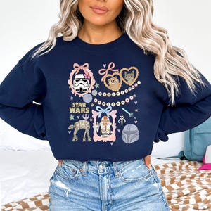 May include: Navy blue Star Wars sweatshirt featuring various character illustrations within pink frames. The design includes a Stormtrooper, Ewoks, R2-D2, and other iconic elements from the Star Wars franchise. The words "STAR WARS" are also printed on the sweatshirt.