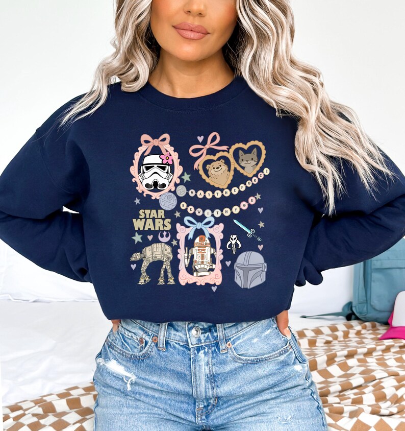 May include: Navy blue Star Wars sweatshirt featuring various character illustrations within pink frames. The design includes a Stormtrooper, Ewoks, R2-D2, and other iconic elements from the Star Wars franchise. The words "STAR WARS" are also printed on the sweatshirt.