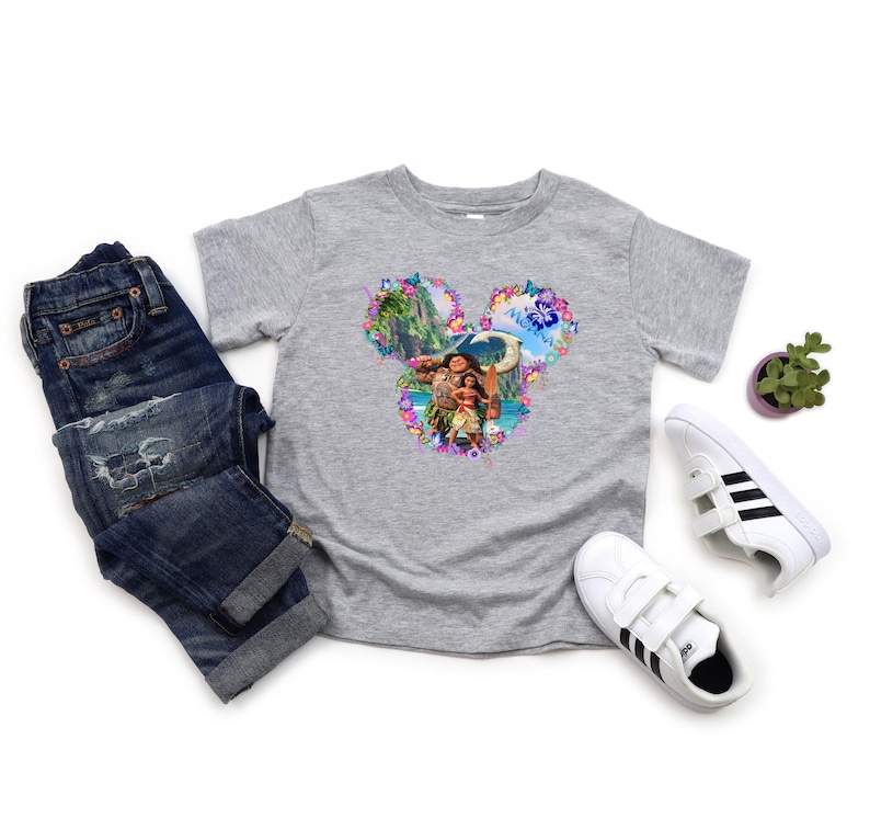 Puede incluir: Una camiseta gris con un gr&aacute;fico colorido de Moana y Maui en forma de cabeza de Mickey Mouse, rodeada de elementos florales. La camiseta se combina con vaqueros azules y zapatillas blancas con rayas negras.