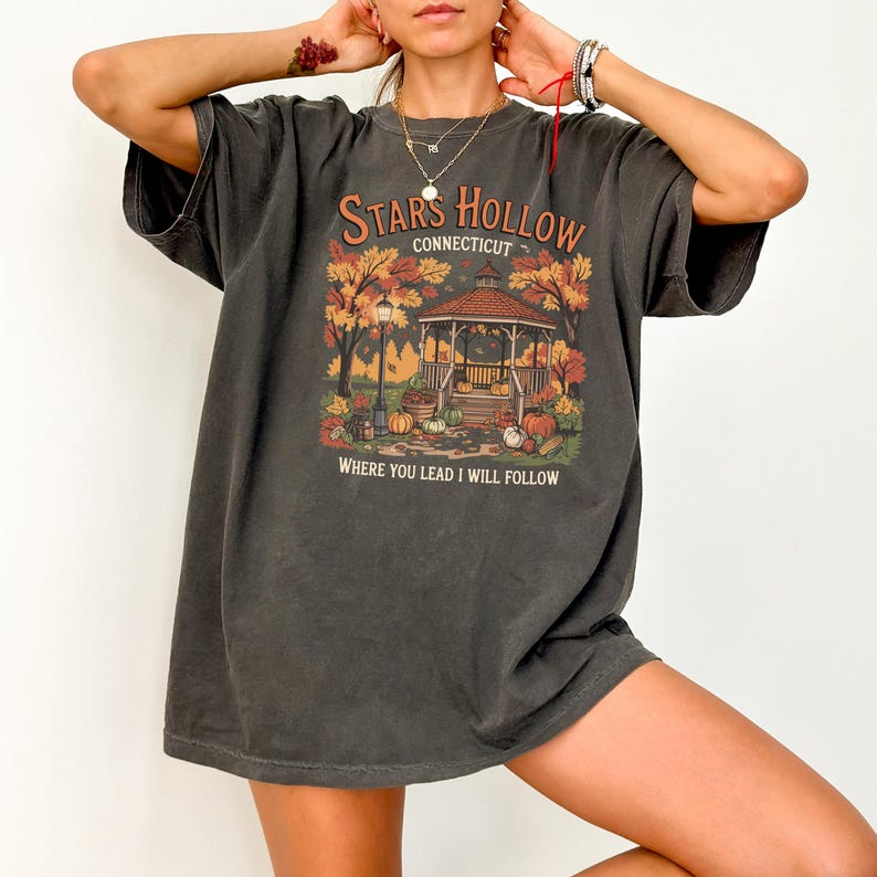 May include: Dark grey t-shirt featuring a fall-themed graphic with a gazebo, pumpkins, and autumn foliage. The text reads "Stars Hollow Connecticut" and "Where you lead I will follow."