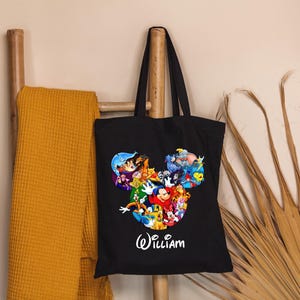 May include: Black tote bag featuring a colorful graphic of Disney characters in a Mickey Mouse head shape. The name "William" is written in white script below the graphic. A yellow blanket hangs on a wooden ladder.