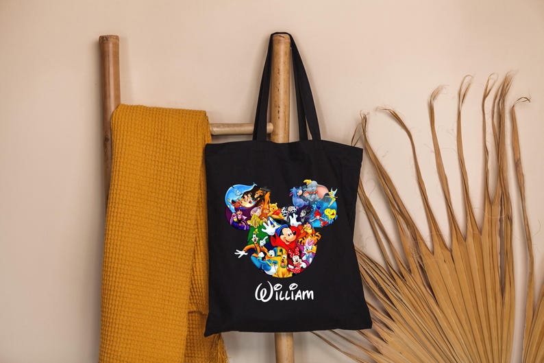 May include: Black tote bag featuring a colorful graphic of Disney characters in a Mickey Mouse head shape. The name "William" is written in white script below the graphic. A yellow blanket hangs on a wooden ladder.