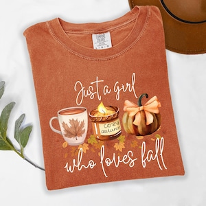 K&ouml;nnte beinhalten: Rostfarbenes T-Shirt mit dem Text "Just a girl who loves fall". Die Grafik zeigt eine Kaffeetasse, eine Kerze und einen K&uuml;rbis. Das Shirt ist von der Marke Comfort Colors, Gr&ouml;&szlig;e L-G.