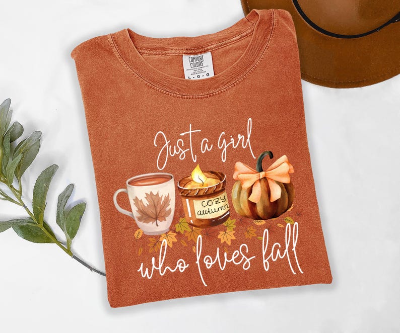 K&ouml;nnte beinhalten: Rostfarbenes T-Shirt mit dem Text "Just a girl who loves fall". Die Grafik zeigt eine Kaffeetasse, eine Kerze und einen K&uuml;rbis. Das Shirt ist von der Marke Comfort Colors, Gr&ouml;&szlig;e L-G.