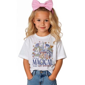 Puede incluir: Camiseta blanca con un gr&aacute;fico colorido que presenta un castillo, flores y el texto "The Most Magical Place on Earth". La camiseta la lleva una persona joven con una diadema de lazo rosa y vaqueros azules.
