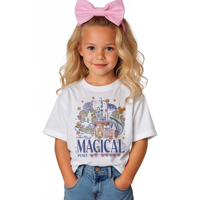 Puede incluir: Camiseta blanca con un gr&aacute;fico colorido que presenta un castillo, flores y el texto "The Most Magical Place on Earth". La camiseta la lleva una persona joven con una diadema de lazo rosa y vaqueros azules.