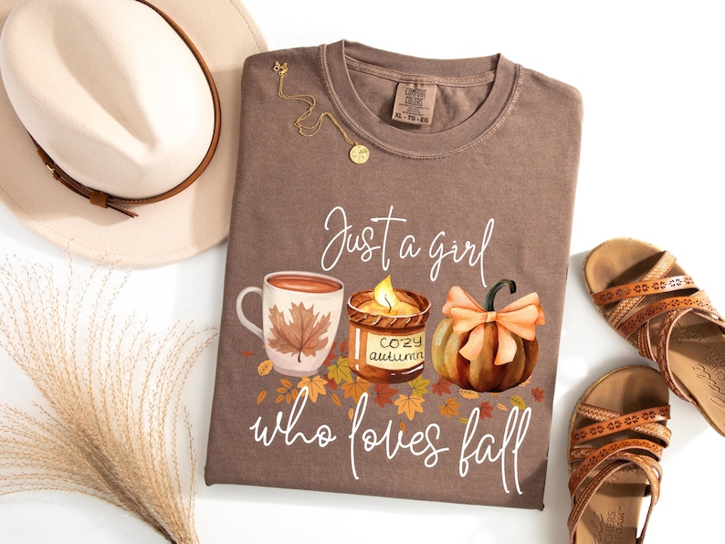 K&ouml;nnte beinhalten: Ein braunes T-Shirt mit dem Text "Just a girl who loves fall" und herbstlichen Grafiken, darunter eine Tasse, eine Kerze und ein K&uuml;rbis. Eine goldene Halskette, Sandalen und ein Hut sind ebenfalls im Bild.
