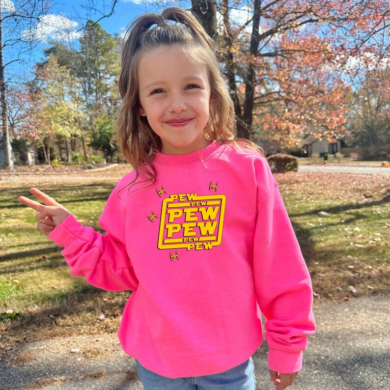 May include: A bright pink sweatshirt with the text "PEW PEW PEW" in yellow and white, framed in a yellow rectangle. The sweatshirt is worn by a person outdoors.