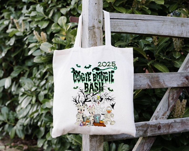 May include: Off-white tote bag with a Halloween-themed graphic. The design features the text "DOGIE BOOGIE BASH 2025" in green, along with cartoon characters dressed as mummies. A spooky tree and bats complete the design.