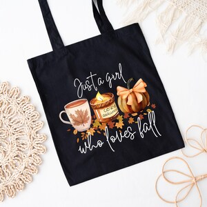 May include: Black tote bag with a fall-themed graphic. The design includes a mug with a leaf, a candle, and a pumpkin, with the text "Just a girl who loves fall."