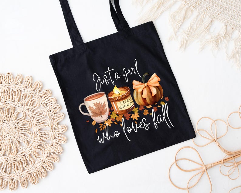 May include: Black tote bag with a fall-themed graphic. The design includes a mug with a leaf, a candle, and a pumpkin, with the text "Just a girl who loves fall."