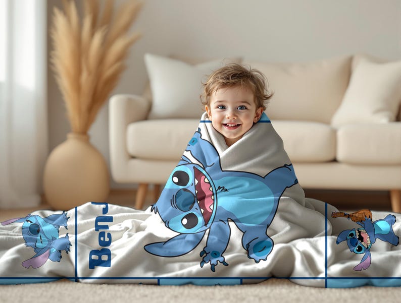 May include: A light gray blanket featuring a large blue cartoon character with big eyes and a wide grin. The blanket has a blue border and the name "Ben" printed on it. A child is wrapped in the blanket.
