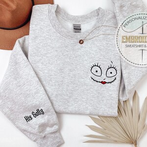 May include: A light gray sweatshirt with a Sally face design on the front and "His Sally" on the sleeve. The design features a black outline with red lips. A brown hat is in the background. The sweatshirt is a personalized gift.