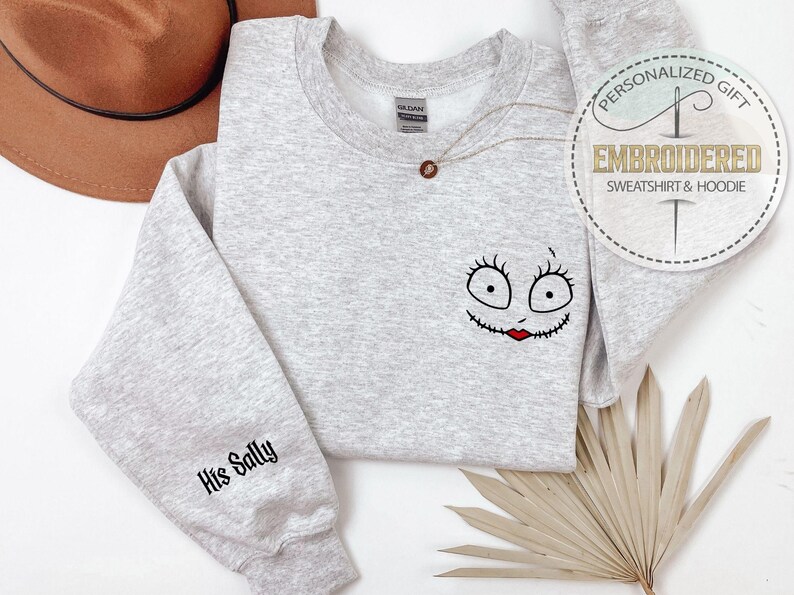 May include: A light gray sweatshirt with a Sally face design on the front and "His Sally" on the sleeve. The design features a black outline with red lips. A brown hat is in the background. The sweatshirt is a personalized gift.